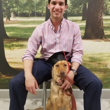 A man in a checkered shirt and navy pants sitting on a stool with a light brown dog sitting between his legs. The dog is wearing a black graduation cap with a red tassel. Both the man and the dog are looking at the camera, with the man smiling.